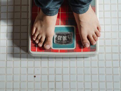 From above crop anonymous barefoot child in jeans standing on weigh scales on tiled floor of bathroom